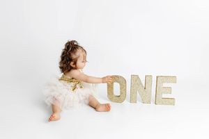 One-year-old baby girl in gold and white tutu dress touching glittery ONE letters on white background.