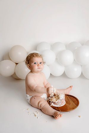 Minimalist cake smash by Menifee photographer - Baby celebrating first birthday covered in cake frosting with white balloon garland.