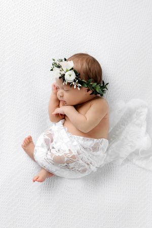 Sleeping newborn baby girl wearing white floral crown wrapped in lace on white textured blanket.