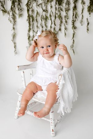 Adorable baby girl in white romper sitting on vintage white rocking chair with floral backdrop.