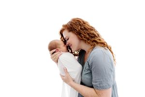 Tender moment of redhead mother gently kissing her swaddled newborn baby against white background.