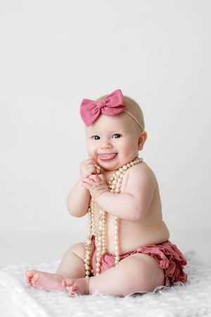 Happy baby girl wearing pink bow headband and ruffled bloomers playing with pearl necklace.