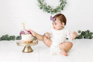 1st birthday cake smash in Temecula - photoshoot of cute baby girl in white lace romper touching her first birthday cake with her foot.
