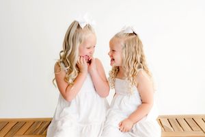 Two little girls in white dresses and hair bows laughing together while sitting on a wooden bench by Temecula photographer.