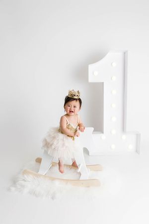 Baby girl in gold sequin dress and crown sitting on rocking horse for 1st birthday photoshoot in Winchester Calilfornia.