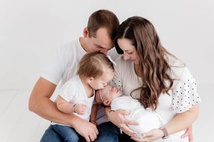 Family of four with parents and toddler lovingly gathered around newborn baby in white studio.