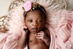 Beautiful newborn baby girl with pink floral headband lying on soft pink feathery blanket.