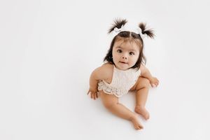 Adorable baby girl with pigtails sitting on white background wearing a cream lace romper.