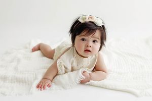 Baby girl in white lace romper with floral headband doing tummy time on white blanket.