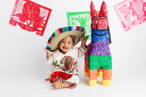 Happy baby in sombrero and traditional Mexican outfit posing with colorful pinata for 1st birthday photoshoot.