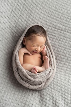 Peaceful smiling newborn baby wrapped in a gray knit blanket on a gray textured background.