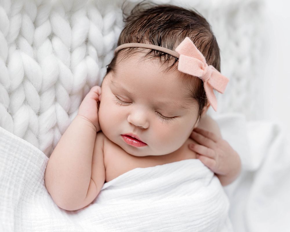 Professional newborn photographer in Menifee portrait of baby girl sleeping peacefully wearing pink bow headband on white knit blanket.