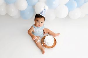 Baby boy in blue outfit with birthday hat sits by smash cake surrounded by blue and white balloons.