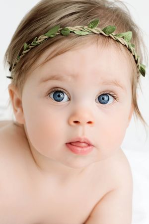 Close-up portrait of a blue-eyed baby wearing a green leaf crown headband looking at camera.