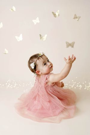 Adorable baby girl in pink tulle dress reaches toward gold butterfly decorations in dreamy photo.