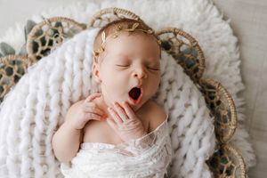 Peaceful newborn baby yawning while swaddled in white wrap with gold leaf headband in basket.