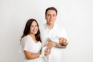 Smiling couple in white outfits proudly holds their newborn baby against a clean white background.