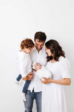 Happy family in white outfits with curly-haired toddler and mother holding newborn baby together.