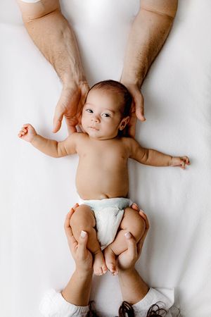 Newborn baby in diaper lying on white blanket while parents gently hold head and feet from above.