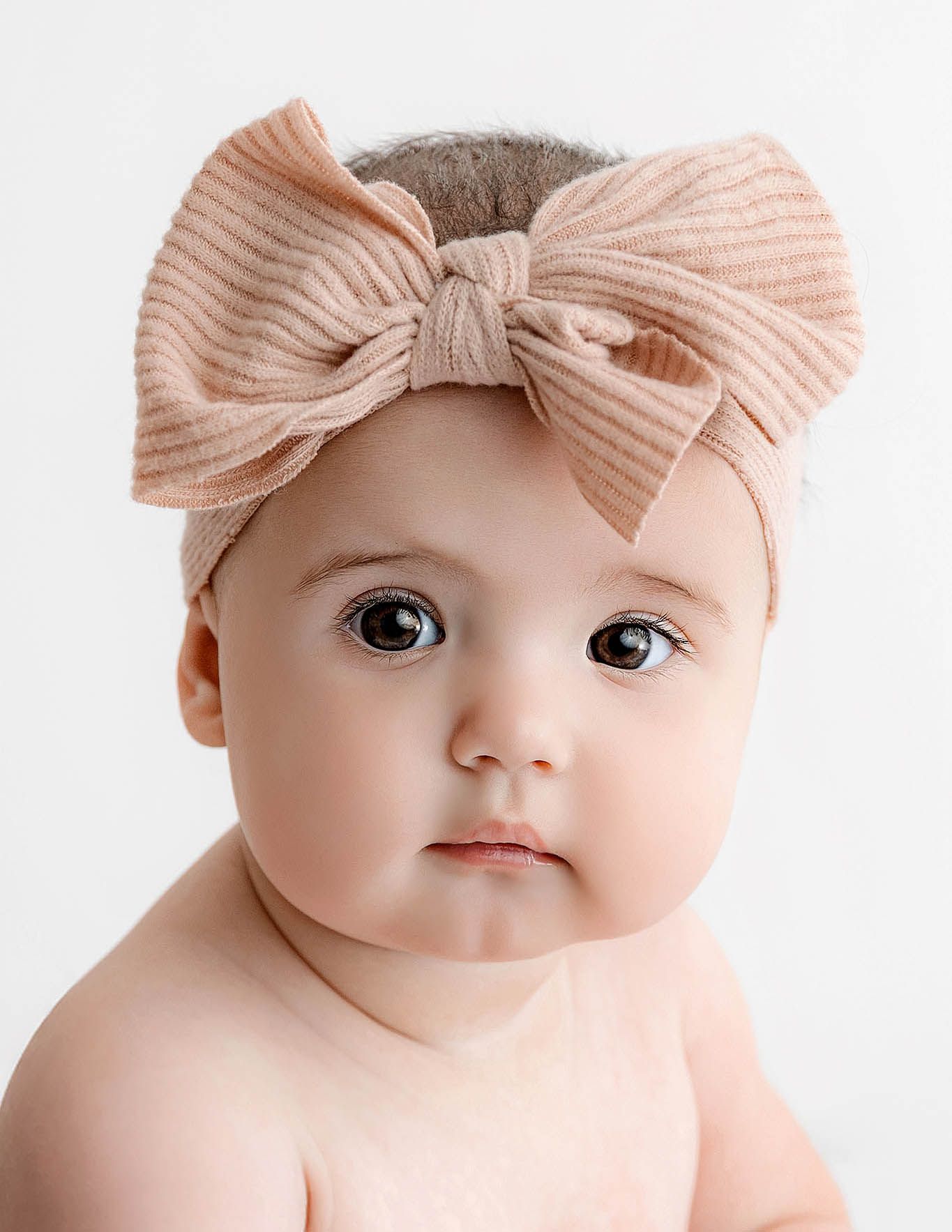 Adorable baby girl wearing a pink ribbed bow headband, with big brown eyes and a sweet expression.