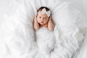 Newborn baby girl with white bow headband lying in soft white blankets looking up at camera.