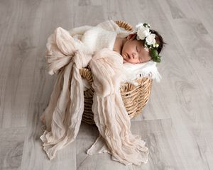 Sleeping newborn baby girl with floral crown nestled in basket with pink bow wrap.