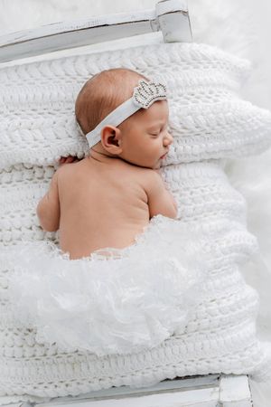 Sleeping newborn baby girl in white ruffled bloomers and crown headband on stacked knit blankets.