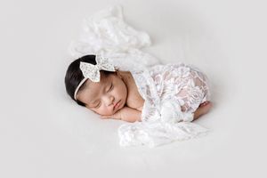Sleeping newborn baby girl in white lace outfit and bow headband on white background.