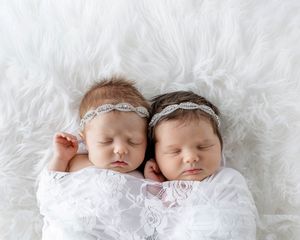 Twin newborn babies sleeping together wrapped in white swaddle with rhinestone headbands.