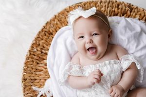 Happy laughing baby girl in white lace dress lying in wicker basket on fluffy white rug.
