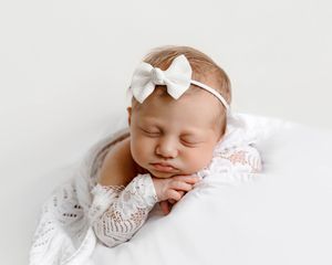 Sleeping newborn baby girl wearing a white bow headband, wrapped in a soft white lace blanket.