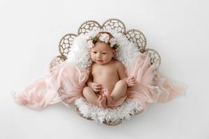 Newborn baby girl resting in a decorative wicker basket with pink tulle and a floral crown on white background.