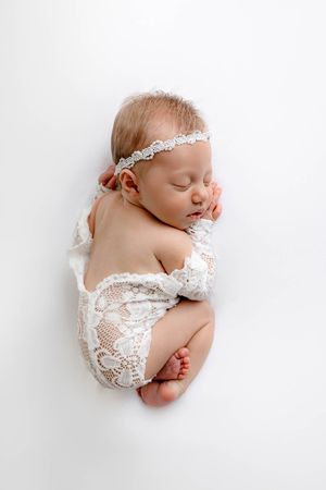 Newborn baby girl in white lace romper and floral headband sleeping peacefully on a white background.