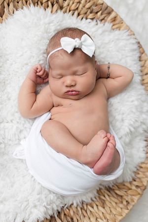Sleeping newborn baby girl with white bow headband wrapped in white swaddle in a wicker basket.