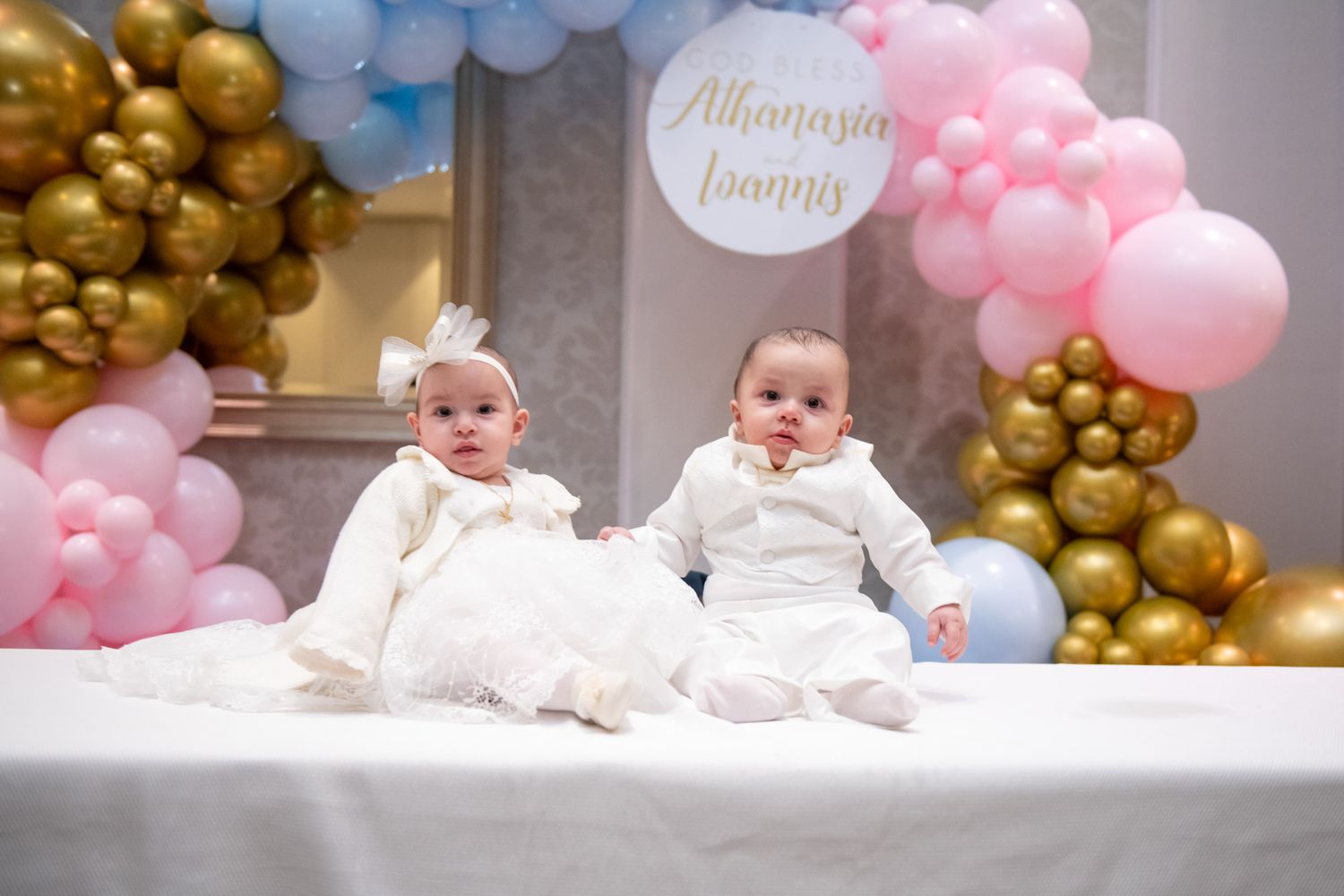 Twin babies in white outfits sit together at a christening party with pink, blue, and gold balloon decor.
