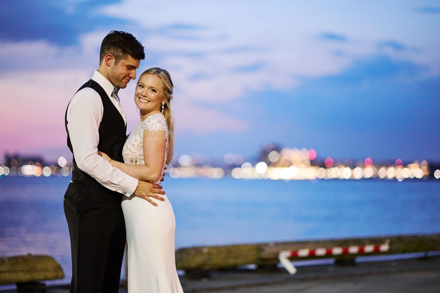 Romantic sunset wedding photos by the water with city lights in background.