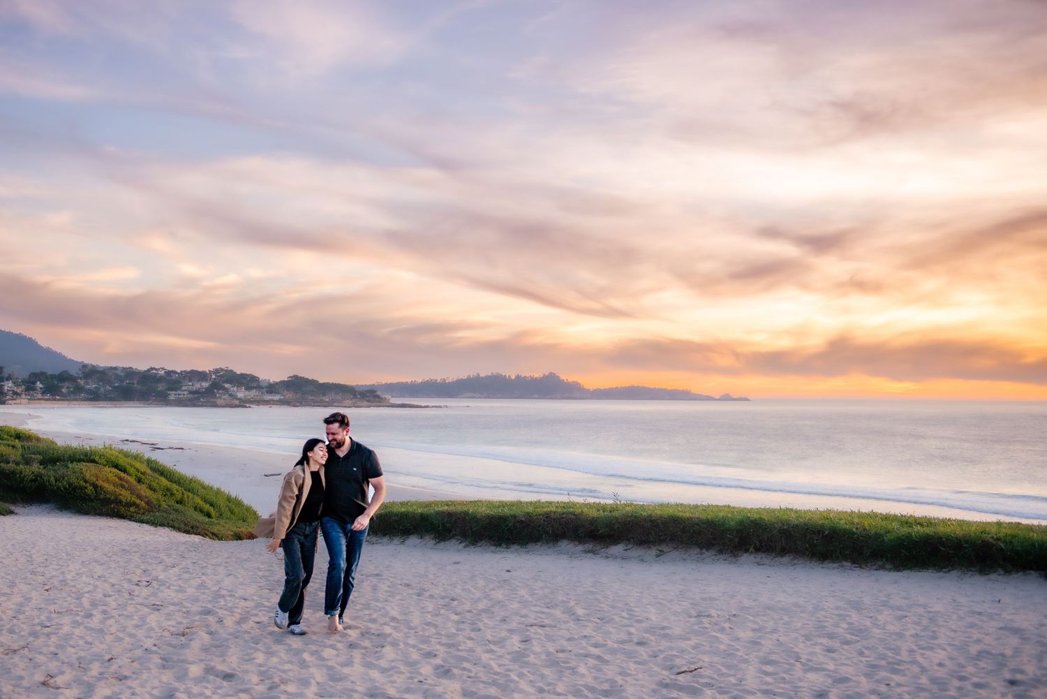 Couple walking on sandy beach at sunset with colorful pink and orange sky, ocean waves, and coastal mountains in background.