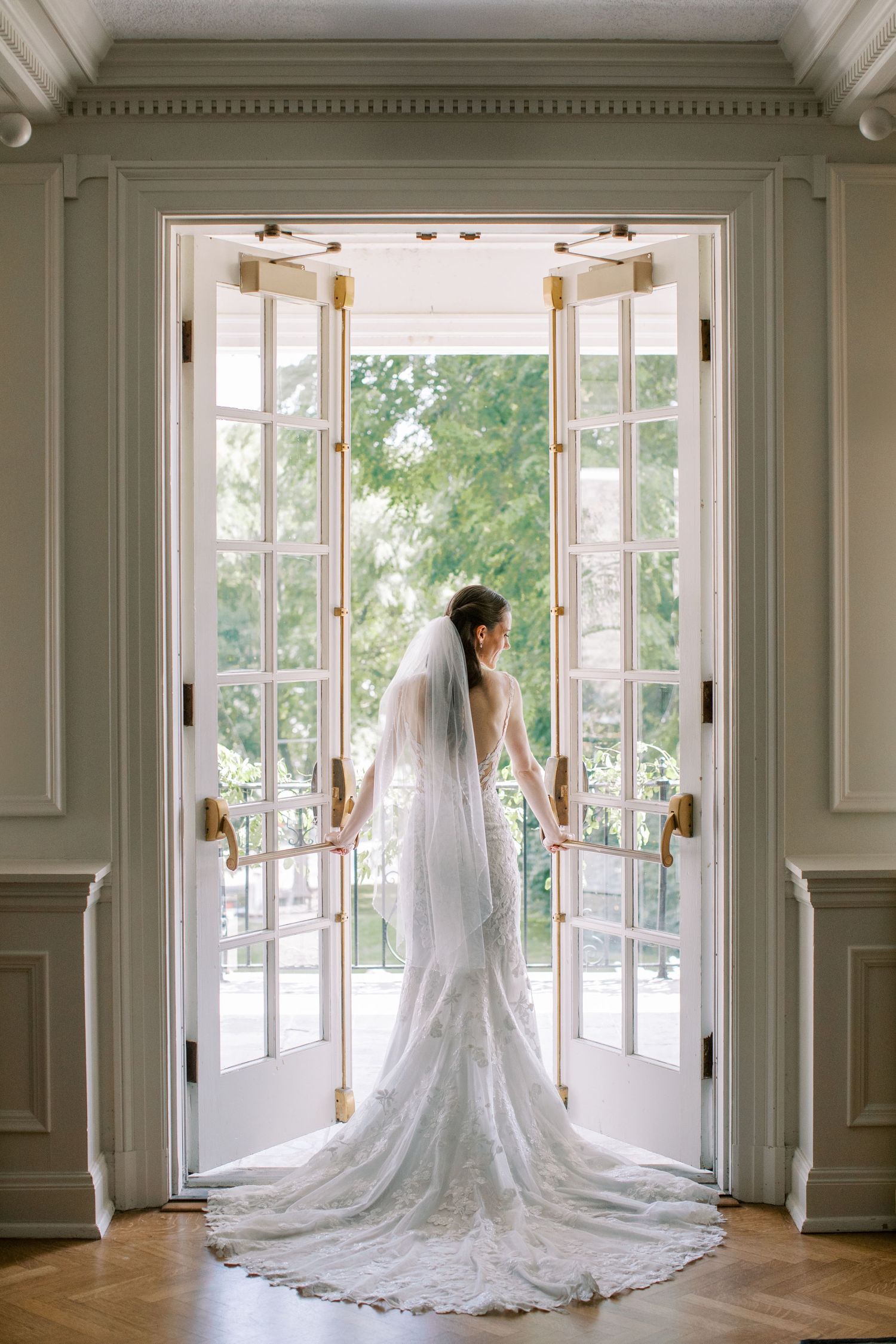 Bride in lace wedding gown and veil stands in open French doors, overlooking lush garden at elegant venue.