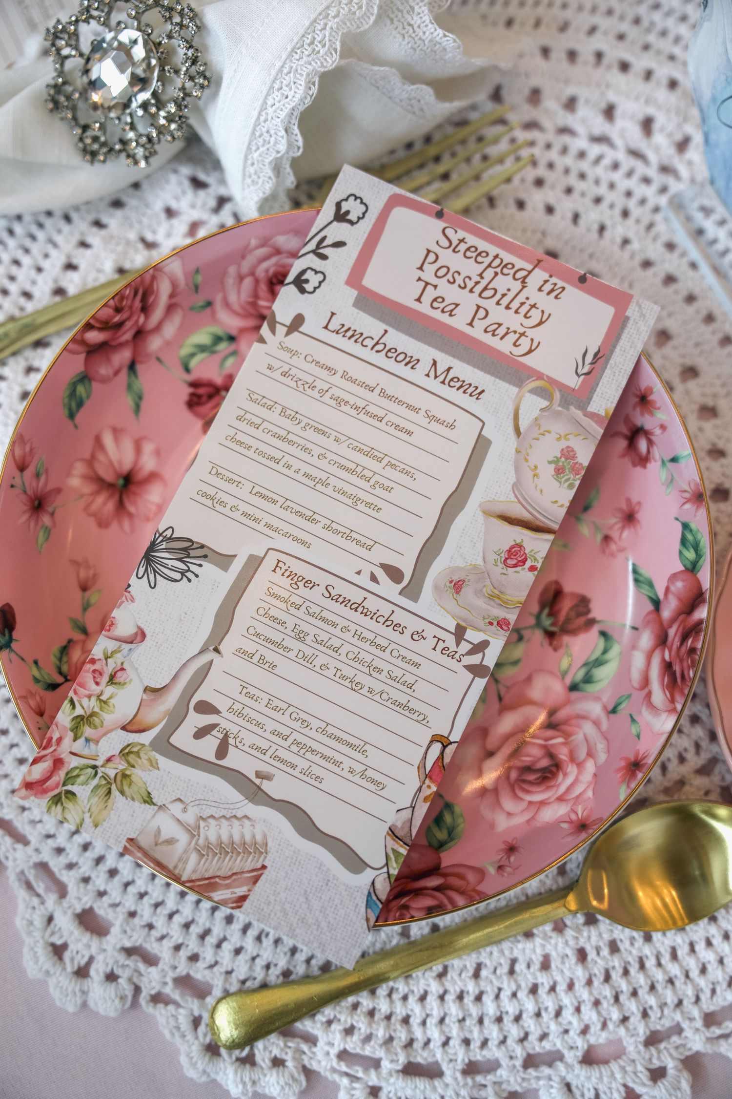 Elegant tea party place setting with floral pink plate, gold spoon, lace doily, and Steeped in Possibility Tea Party luncheon menu card.
