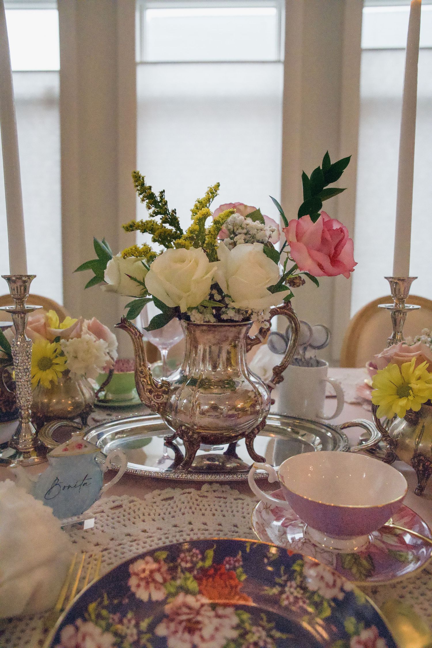 Elegant tea party table with silver teapot floral centerpiece, pink roses, candles, floral teacups, and lace tablecloth.