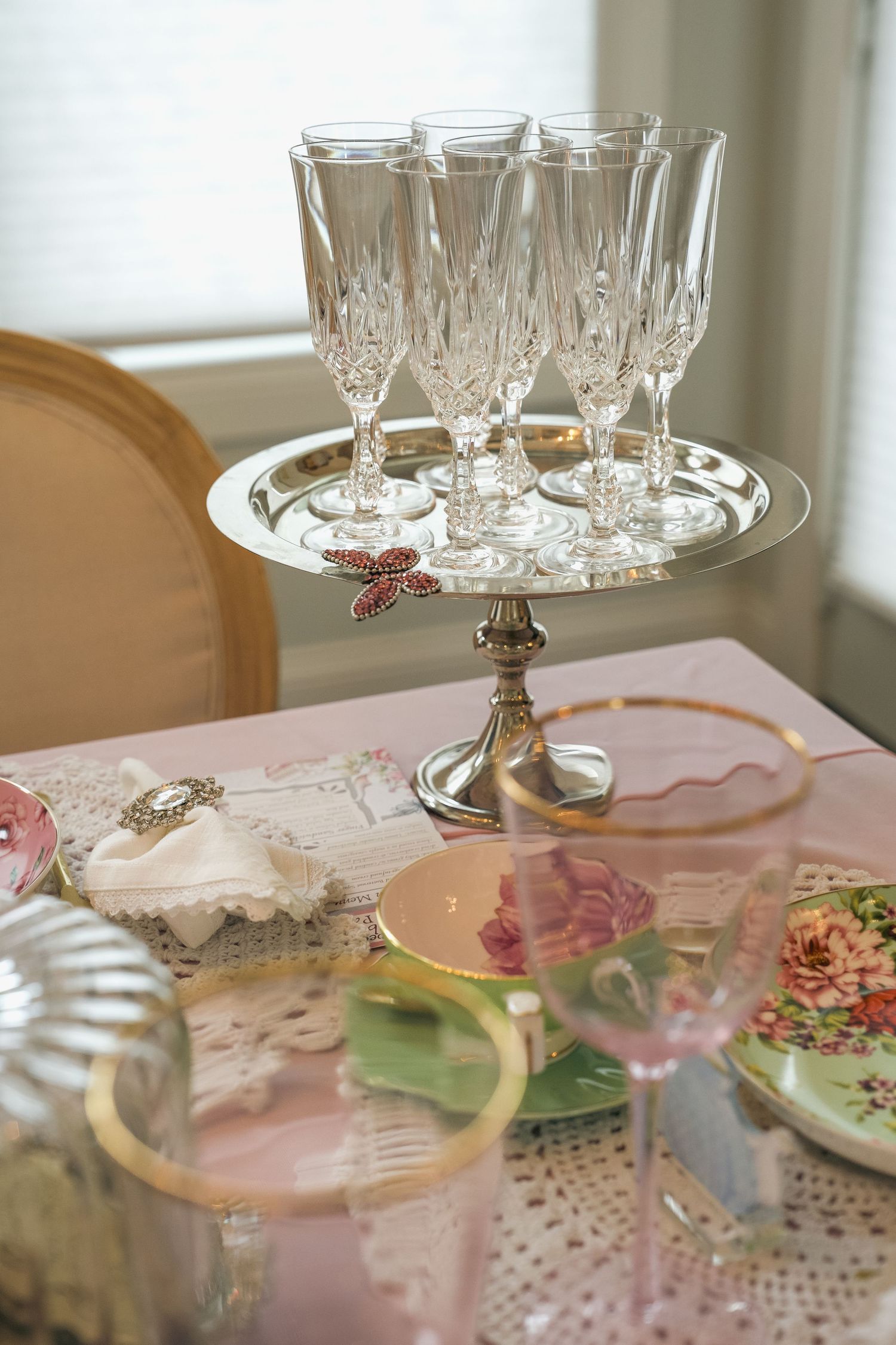 Elegant table setting with crystal champagne flutes on silver cake stand, floral china plates, and pink tablecloth for a tea party.