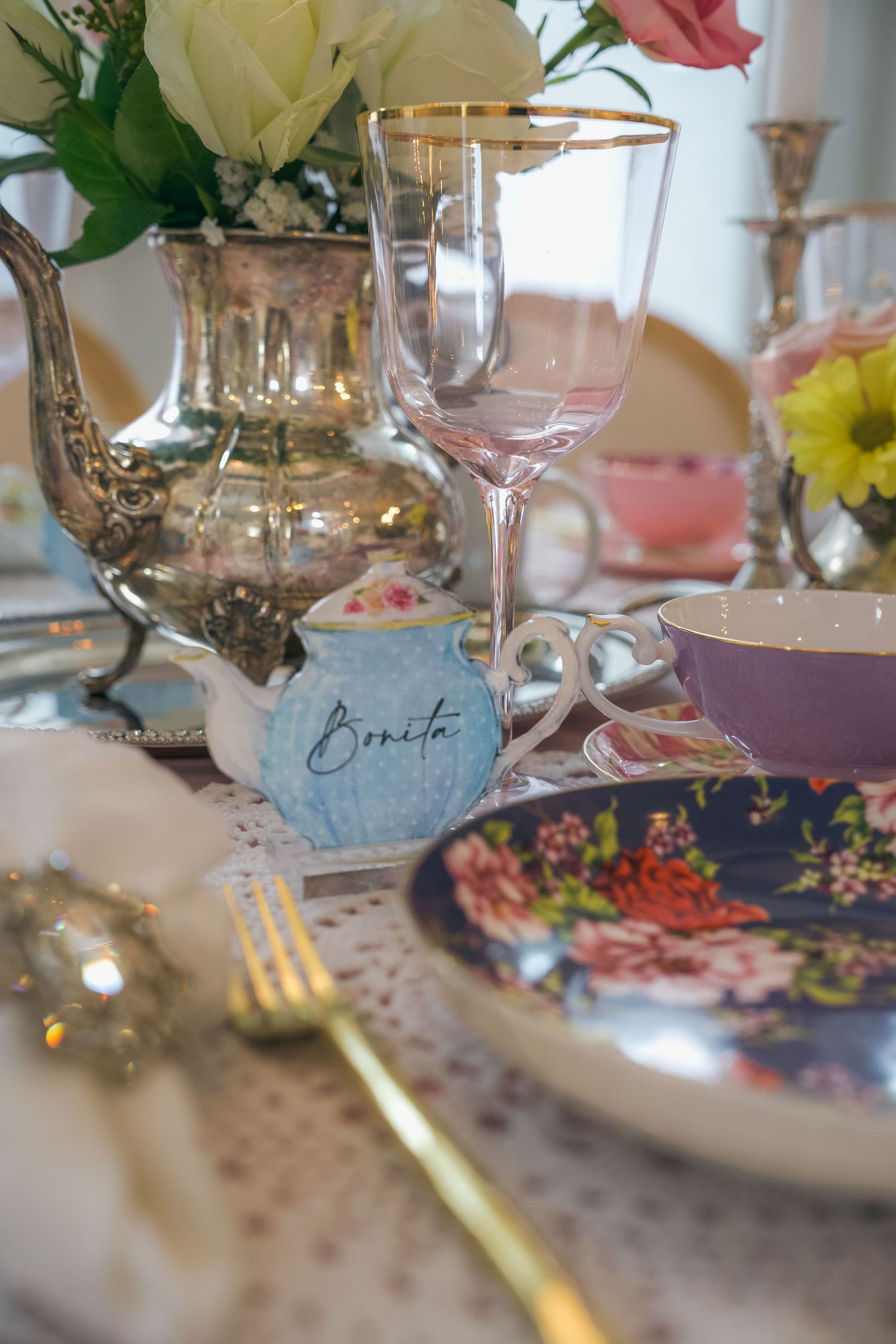 Elegant tea party table setting with floral china, gold fork, crystal wine glass, silver teapot, and a small blue teapot labeled Bonita.