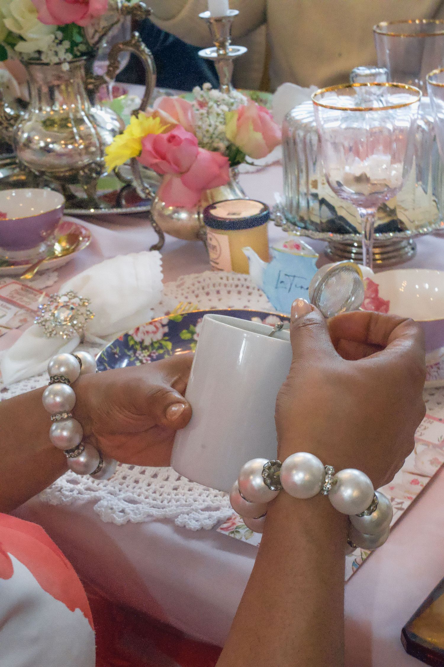 Elegant tea party table with pearl bracelets, silver teapot, floral cups, pink roses, and crystal glasses on lace tablecloth.