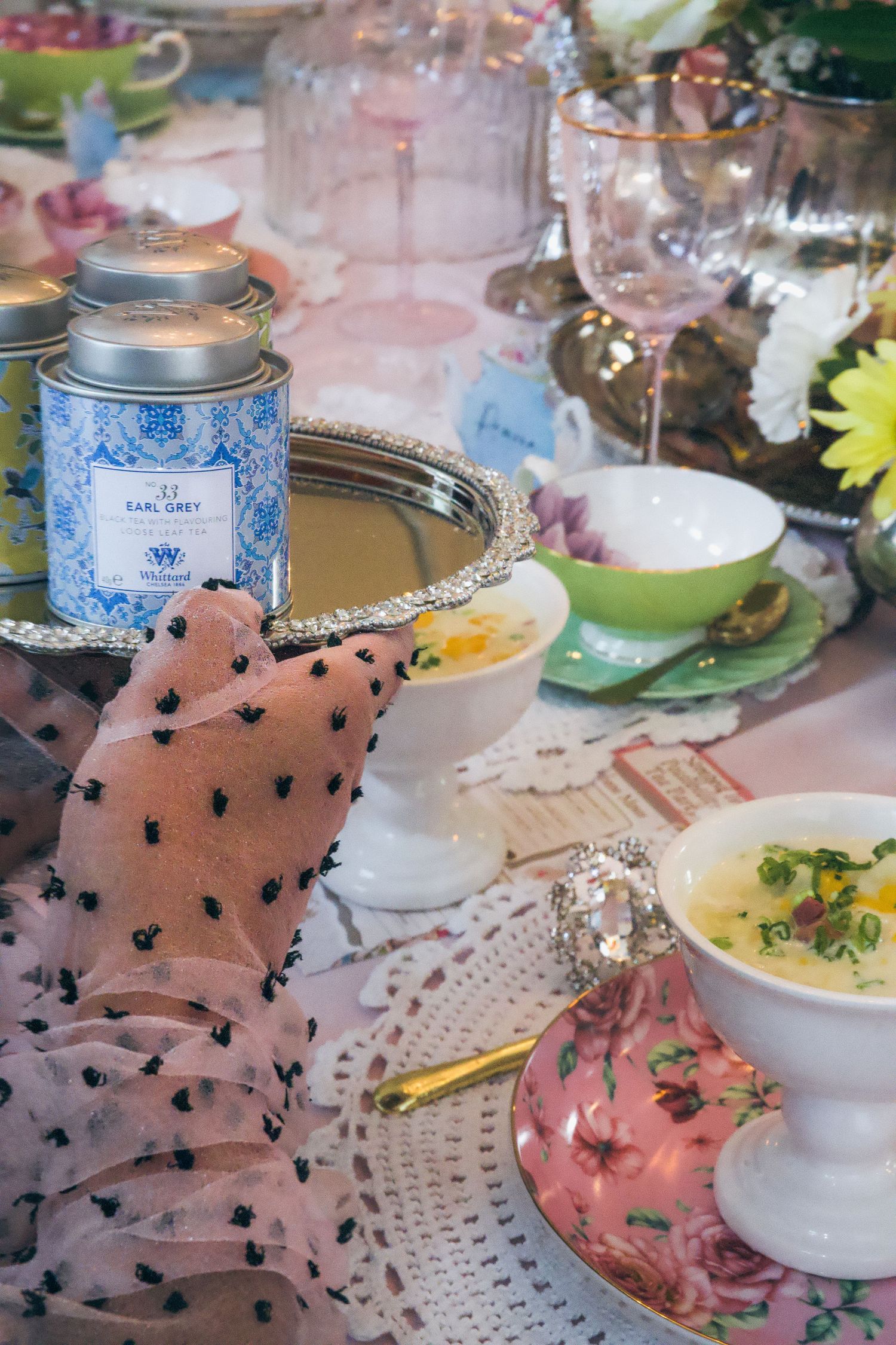Elegant tea party table with Earl Grey tin, floral teacups, soup bowls, and wine glasses on a lace tablecloth.