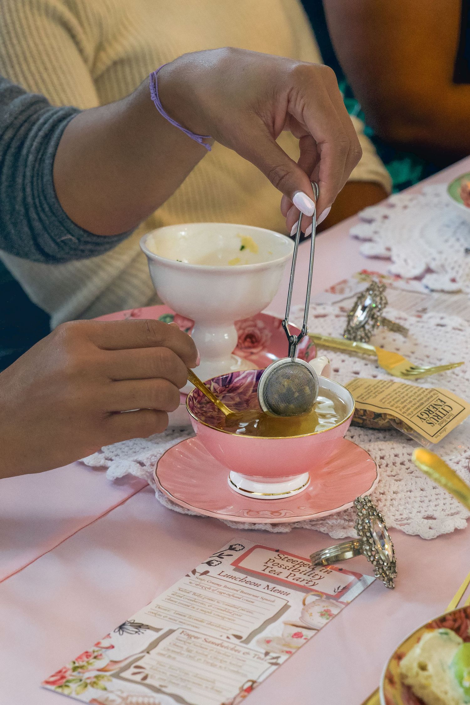 Woman steeping loose leaf tea in a pink teacup with a strainer at an elegant tea party table.