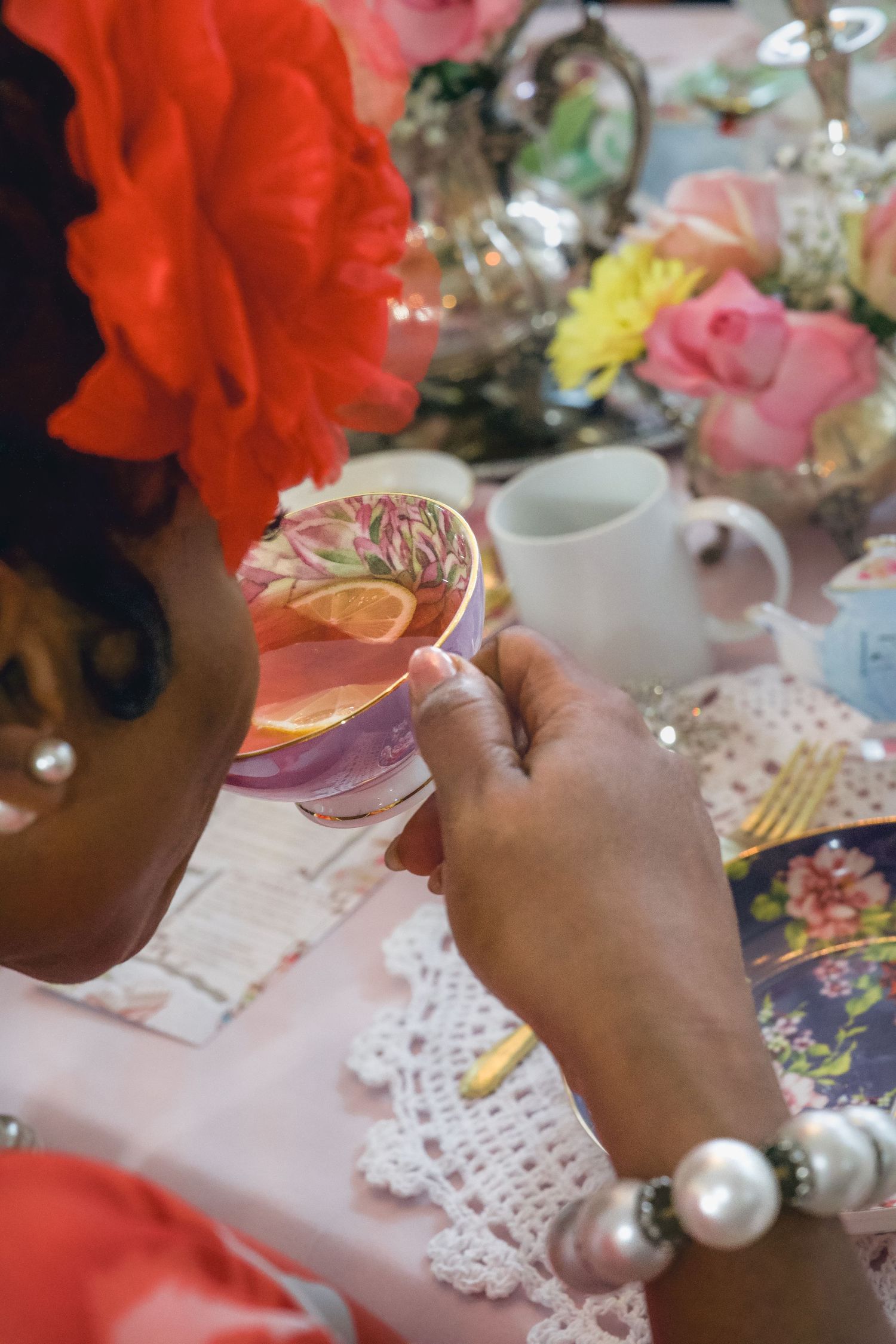 Woman wearing pearl bracelet and red flower holds floral teacup at elegant tea party table with roses and silver teapot.