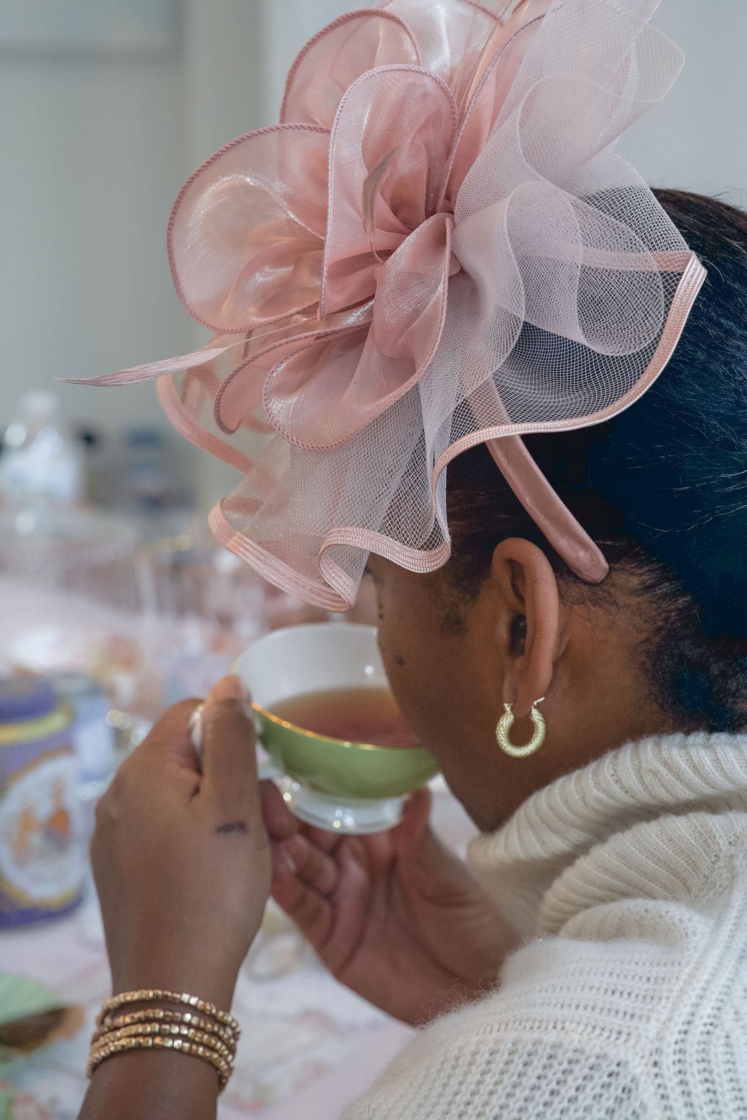 Elegant woman wearing pink fascinator hat sipping tea from green cup at formal tea party.