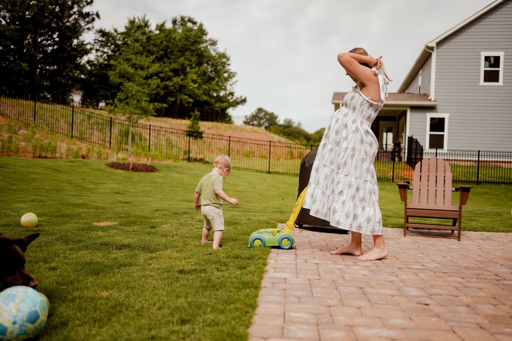 Woman in floral dress plays with toddler pushing toy mower in sunny backyard near patio.