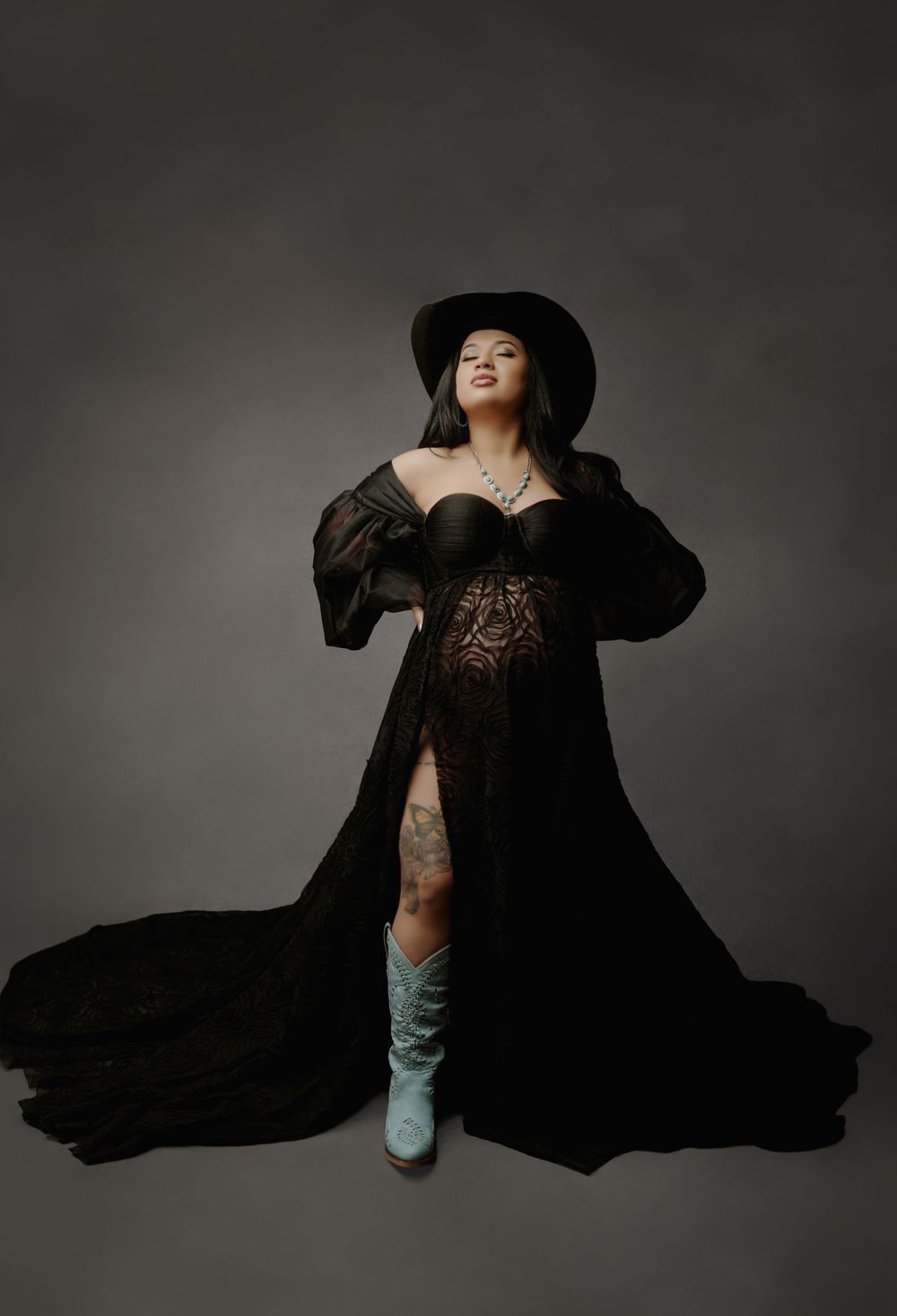 Pregnant woman in black lace maternity gown, wide-brim hat, and turquoise cowboy boots poses elegantly in studio.