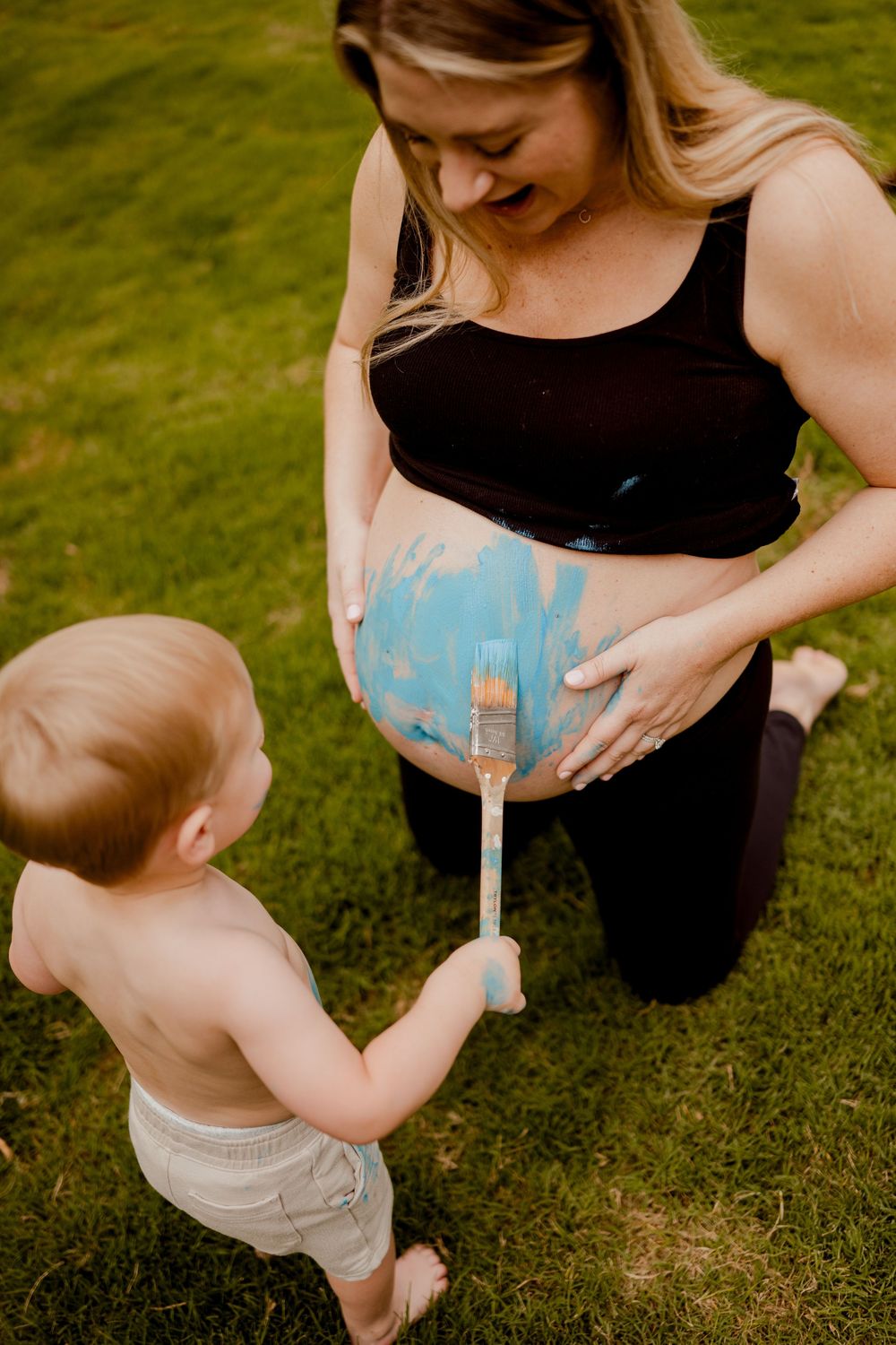 Toddler painting pregnant mother's belly with blue paint outdoors on grass during a gender reveal maternity photo shoot.